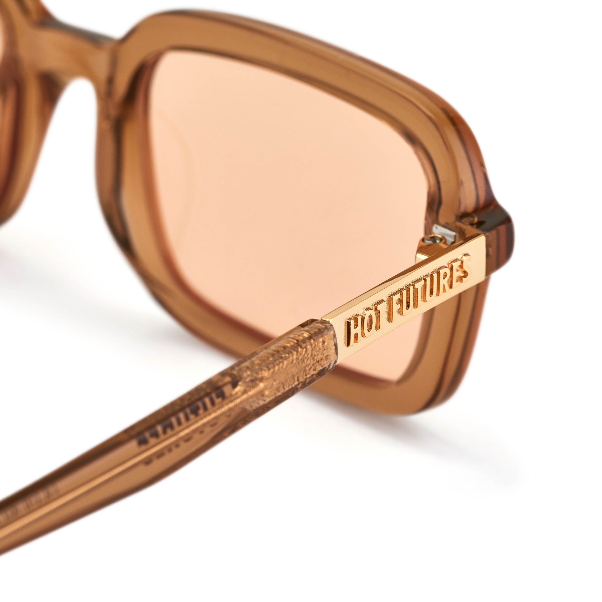 A pair of beige rectangular sunglasses with amber tinted lenses. Shot close up showing their lens colour and gold branding text logo on sides